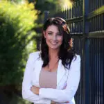 Confident woman in white blazer standing outdoors by a fence.