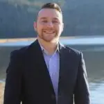 A smiling man in a suit stands outdoors near water with a blurred background.