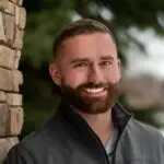 Smiling bearded man outdoors near brick wall.