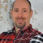 Smiling man in a red plaid shirt with a light gray background.