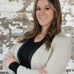 Confident woman in business attire smiling with folded arms against a textured wall.