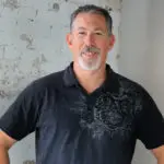 Smiling man in a black polo shirt standing indoors.