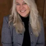 Woman with long blonde hair smiling, wearing a gray jacket and black top.
