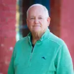 Elderly man in a teal pullover stands against a red brick wall.