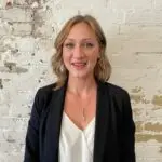 Smiling woman in business casual attire against a textured white brick wall.