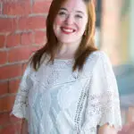 Smiling woman in white lace top standing by a brick wall.