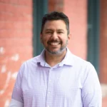 Smiling man in a light purple shirt standing outdoors.