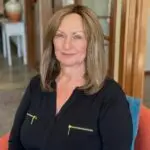 A smiling woman with shoulder-length blonde hair sitting indoors.