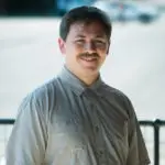 A man with a mustache smiling outdoors in a casual shirt.
