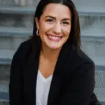 Smiling woman in black blazer sitting on stairs.
