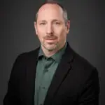 Professional man in black blazer and green shirt against dark background.